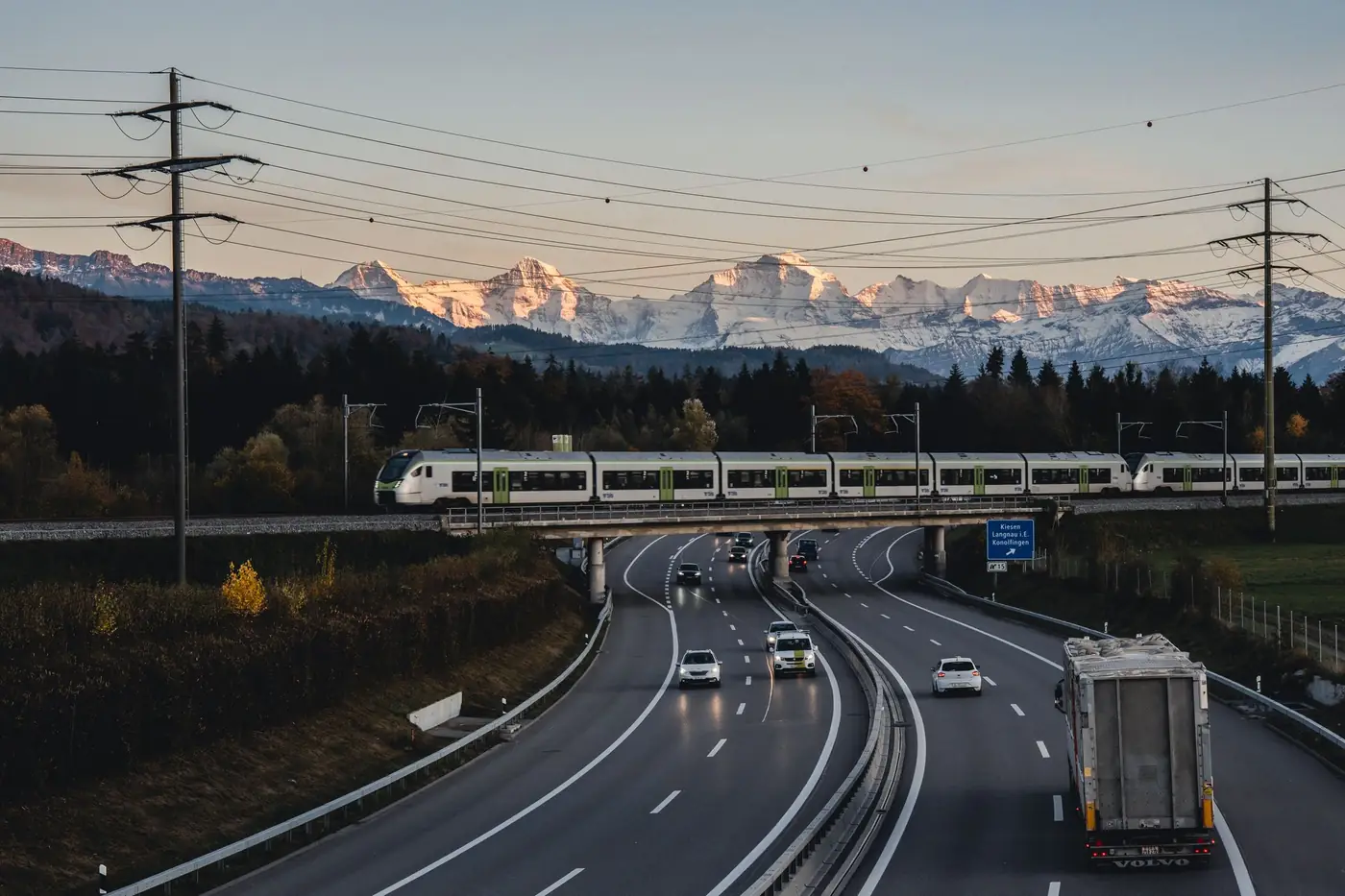 Trein die bij zonsondergang over een brug boven een snelweg in Zwitserland rijdt, met bergen op de achtergrond