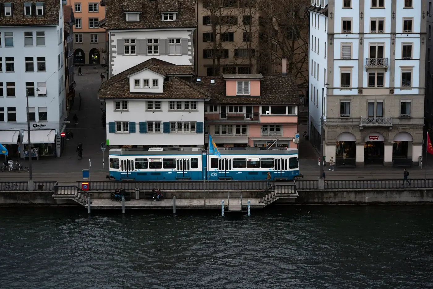 A blue tram travels along the riverbank in Zürich, showcasing local architecture.