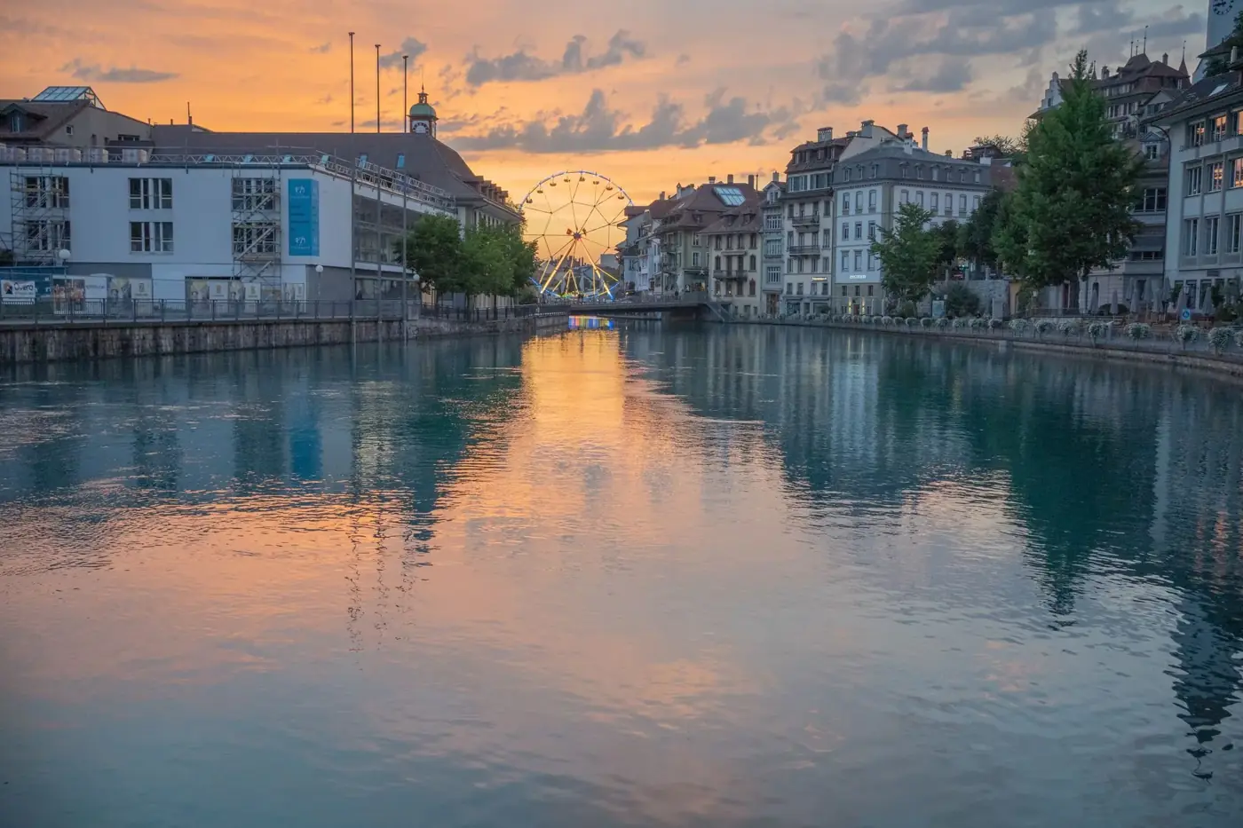 Beautiful sunset view of Thun, Switzerland, with a Ferris wheel reflected in the river.